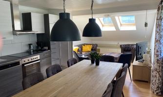 A modern dining room with a wooden table and black pendant lights. In the background, a cozy seating area and windows with daylight can be seen.
