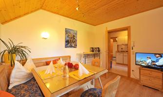 A cozy living area with a wooden ceiling and a table with chairs. In the background, an open kitchen and a television are visible.
