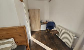 A simple room with a bed, a desk, and a chair. The walls are white and there is a radiator.