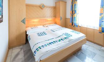A cozy bedroom with a double bed and wooden paneling. The room has a window with colorful curtains and a bright tiled floor.