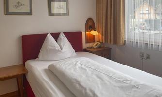 A simple room with a single bed and white sheets. A nightstand and a window with curtains complete the furnishings.