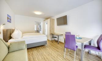 A modern room with a double bed, a desk, and a television. The decor is cozy and bright, with a large window and a sofa.