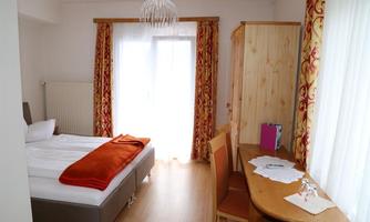 A bright room with a single bed, a desk, and chairs. The large windows let in plenty of natural light, and the curtains are designed in vibrant colors.