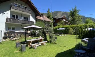 A cozy garden with plenty of greenery and various seating options. In the background, houses and mountains are visible.
