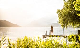 A person stands at the shore of a quiet lake and looks into the distance. The surroundings are filled with lush greenery and gentle hills, while the sun shines softly.