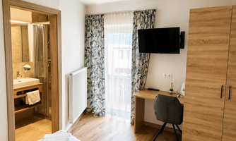 A modern hotel room with a light wood floor and a large window. There is an en suite bathroom, a desk, and a television.