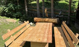 A wooden terrace table with benches in a green, forested area. The furniture is made of solid wood and is located on a lawn.