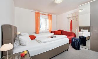 A modern hotel room with two beds and a cozy red sofa. The windows are decorated with orange curtains and there is a suitcase and fresh fruit on the table.