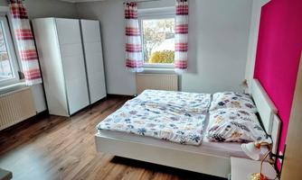 A modern bedroom with a double bed and a wardrobe. The walls are painted light and there are large windows with red curtains.