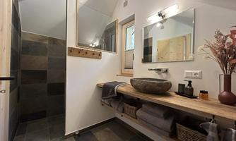 A modern bathroom with an open shower and an elegant stone sink. The wooden shelves are decorated with towels and a beautiful flower vase.