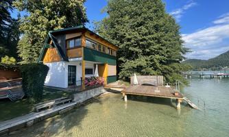 A beautiful house by the water with a dock and surrounded by trees. The clear water surface reflects the blue sky.