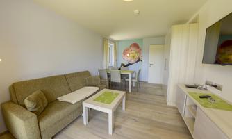 A cozy living room with a green sofa and a round table. A dining area with chairs and a colorful wall decoration is also visible.