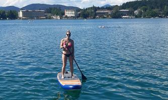 A person is standing on a stand-up paddle board on calm water. The sky is clear and blue with a few clouds.