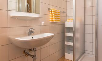A modern bathroom with a shower, a sink, and bright tiles. Towels hang on the wall and a yellow rug lies on the floor.