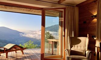 A cozy room with a view of mountains and clouds. The large window front opens up to a terrace with a lounge chair.