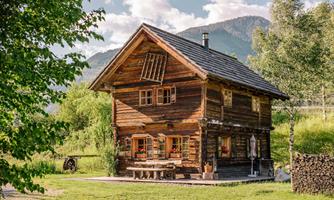 A charming wooden house surrounded by green meadows and trees. In the background, gentle hills can be seen.