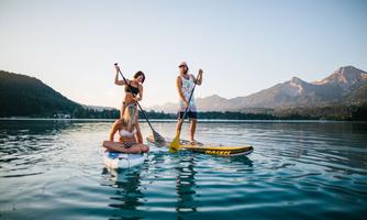 Three people are standing on stand-up paddleboards on a calm lake. In the background, mountains and a clear sky can be seen.