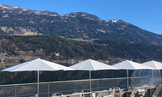 A terrace by the water with loungers and parasols. In the background, mountains and a clear blue sky are visible.
