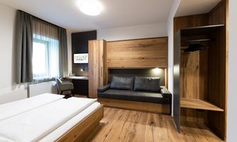 A modern hotel room with wood paneling and a cozy sofa bed. Large windows ensure plenty of daylight and an inviting atmosphere.