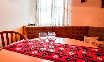 A cozy dining area with a round table and two glasses. The tablecloth is red with a decorative pattern, and there is a large window with curtains.
