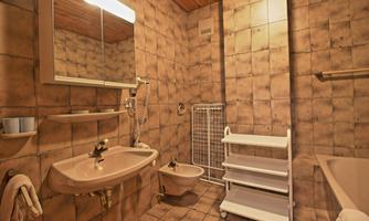 A simple bathroom with tiled walls and a wooden ceiling. It contains a sink, bidet, bathtub, and an open shelf.