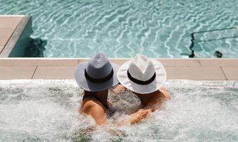 Two people with hats are relaxing in the hot tub. In the background, a calm pool can be seen.