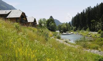 An idyllic landscape with wooden houses next to a tranquil river. Surrounded by green meadows and wooded hills.