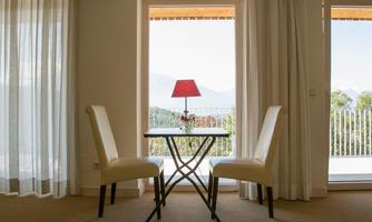 A cozy table with two chairs and a table lamp is situated in front of a window overlooking the countryside. The curtains let in soft light.