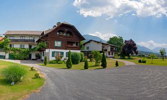 A picturesque country house with large windows and a well-kept garden. The surroundings are green and peaceful, surrounded by gentle hills and a clear sky.