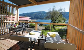 A cozy balcony with two loungers and a small table. In the background, a clear lake and the surrounding mountains can be seen.