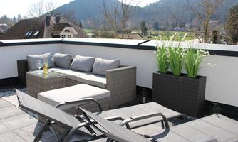 A modern rooftop terrace with comfortable furniture and green plants. In the background, hills and a clear sky can be seen.