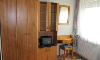 A simple room with a wooden wardrobe, a small desk, and a television. The room is bright and has a large window with curtains.