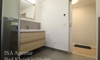 A modern bathroom with a wooden vanity and a large mirror. The walls are light and the floor is gray tiled.