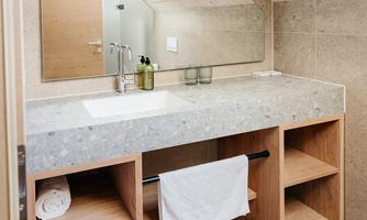 A modern bathroom with a large stone sink and a wooden shelf. The ceiling is slanted, and there is a large mirror above the sink.