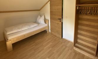 A simple room with a single bed and wooden symbols. In the background, a door and a staircase are visible.