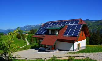 A modern house with solar panels on the roof. The surroundings are green with mountains in the background and a clear, blue sky.