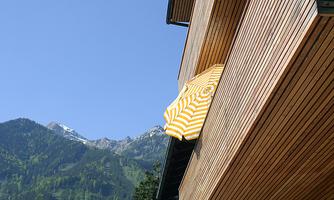 A modern wooden building with a yellow sun umbrella extending outwards. In the background, there are green mountains and a blue sky.