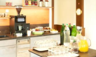 A modern kitchen with a coffee machine and a table full of drinks. There are glasses, bottles, fruit, and small snacks.