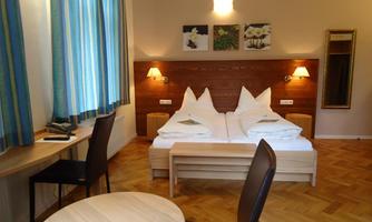 A cozy hotel room with two single beds and wooden flooring. Striped curtains hang at the window, and there is a small table with chairs.