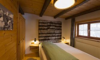A cozy bedroom with a wooden ceiling and a large window. Two single beds with green pillows and soft lighting.