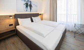 A modern hotel room with a large double bed and bright curtains. On the wall hangs a picture of skiers in a winter landscape.
