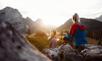Two people are sitting on a rock and looking at the snow-covered mountains. The sun is rising on the horizon, creating a beautiful evening mood.