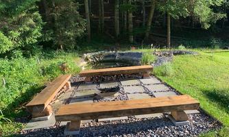 A beautiful outdoor space with wooden benches and a fire pit. Surrounded by green countryside and forest.