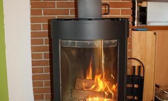 A modern fireplace stove with a clear viewing window through which the fire can be seen. The stove is situated in a cozy room with wooden flooring and red bricks.