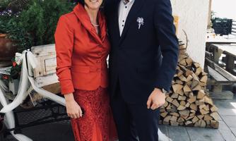 A couple is posing in elegant clothing in front of a cozy backdrop. Behind them, a woodpile and a decorative setting can be seen.
