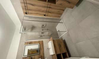 A modern bathroom with a glass shower and wooden shelves. The walls are gray and the floor is tile-like.