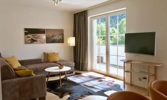 A modern living room with a gray sofa and a round table. Large windows let in plenty of daylight and offer a view of greenery.