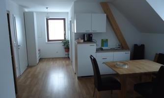 A bright, modern apartment with a small kitchen and a dining area. The room has large windows and wooden flooring.