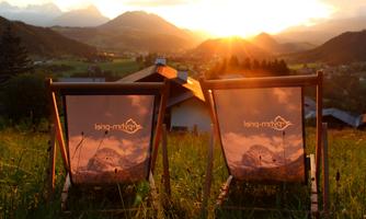 Two deck chairs are set on a meadow with a view of the mountains. The sun is setting behind the mountains, creating a warm atmosphere.