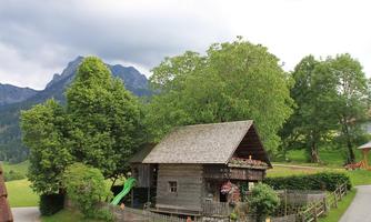 A charming wooden house surrounded by trees and a green garden. In the background, mountains and a sky with clouds can be seen.
